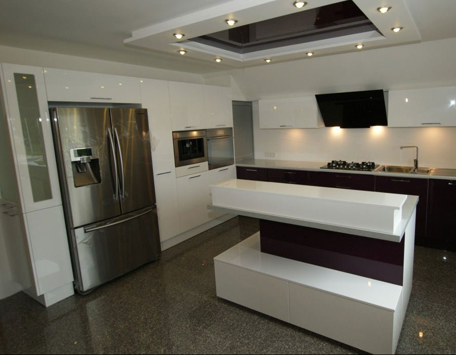 Nowoczesna kuchnia z białymi, lakierowanymi frontami, wyspą kuchenną z blatem w kształcie litery L i burgundowymi akcentami, oraz dużym, dwudrzwiowym, srebrnym lodówką.