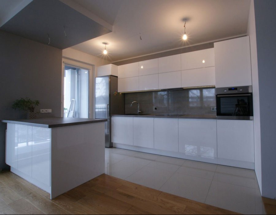 Biała, nowoczesna kuchnia z wyspą, połyskującymi frontami i szarym blatem, oświetlona lampami sufitowymi, z widocznym oknem i drewnianą podłogą w dolnej części kadru.