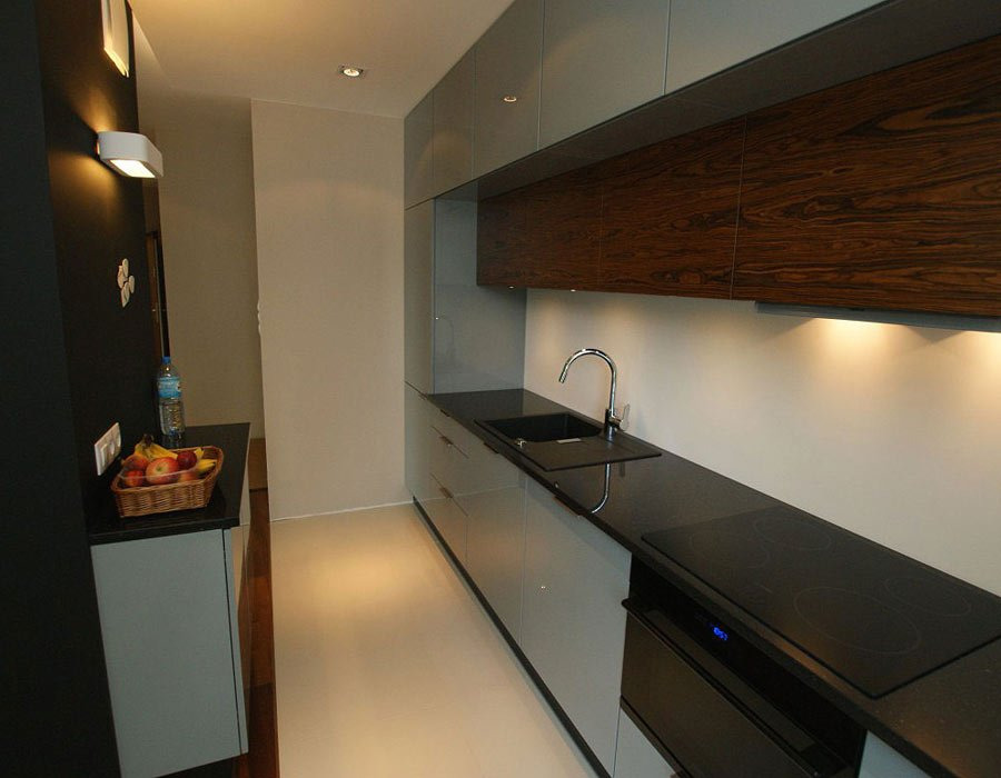 Nowoczesna kuchnia z szarymi szafkami, blatem z czarnego kamienia, drewnianymi szafkami wiszącymi i oświetleniem podszafkowym, z koszykiem owoców na blacie.