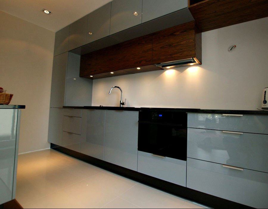 Nowoczesna kuchnia z szarymi, lakierowanymi frontami, ciemnym blatem i drewnianymi szafkami wiszącymi z oświetleniem punktowym nad płytą grzewczą i piekarnikiem w zabudowie.