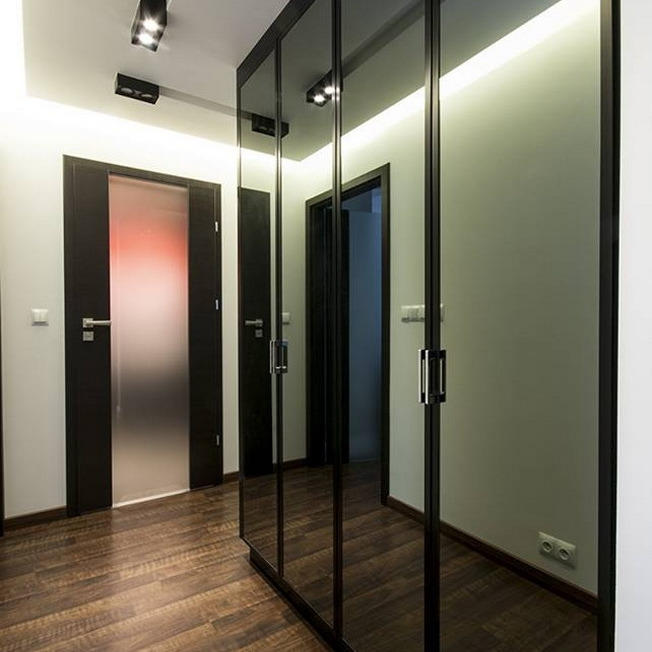 Nowoczesna szafa wnękowa z czarnymi, lustrzanymi drzwiami przesuwnymi, oświetlona listwą LED, widoczna w korytarzu z drewnianą podłogą i drzwiami w ciemnej ramie.