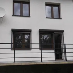 Czarna, pozioma balustrada balkonowa z metalu na tle białej elewacji budynku z brązowymi oknami i satelitą. Widok z dołu, lekko z prawej strony.