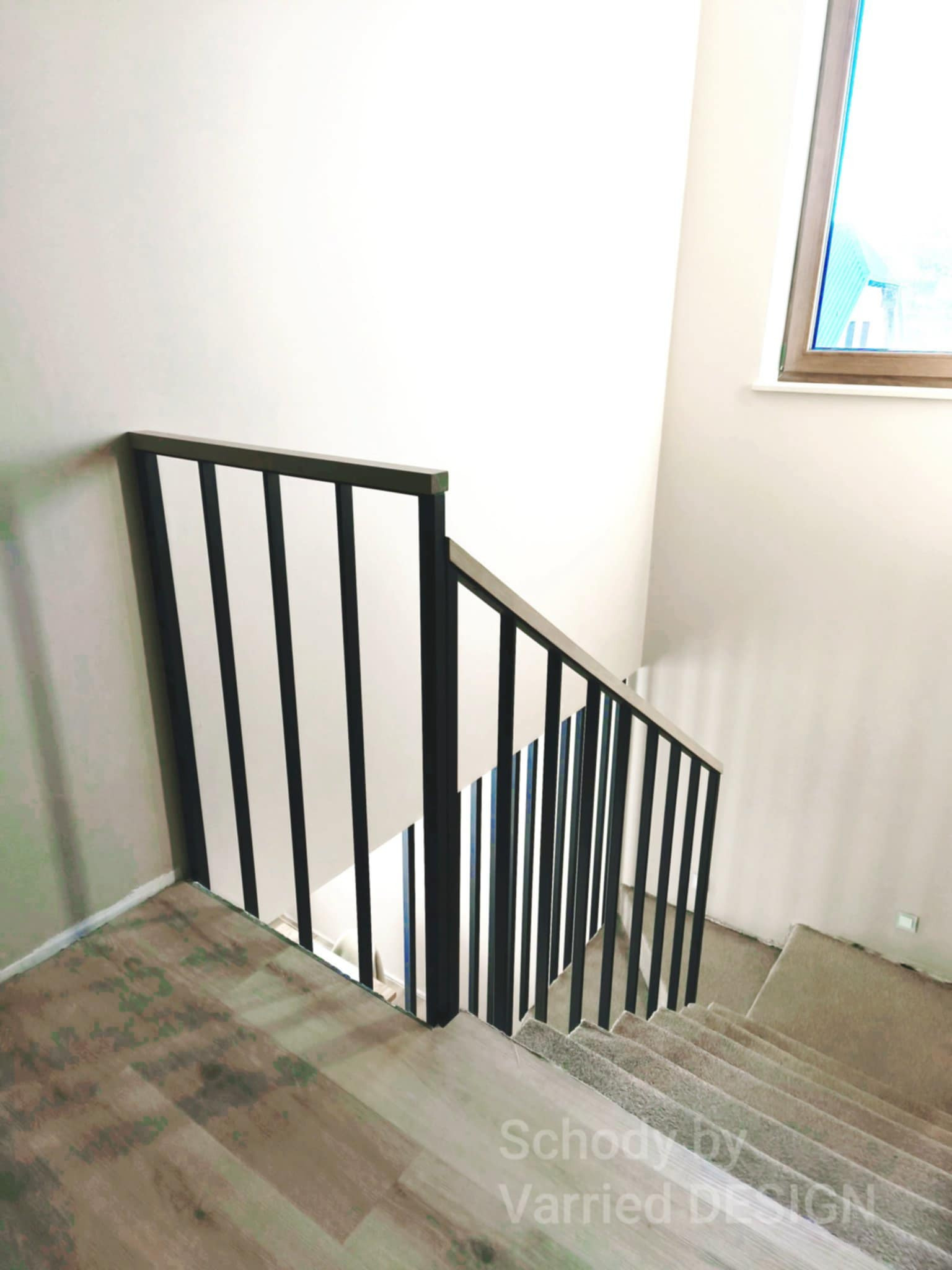 Minimalistyczne schody z czarną, pionową balustradą i drewnianym wykończeniem, widziane z góry. Jasne ściany i okno w tle. Podłoga wykończona panelami.