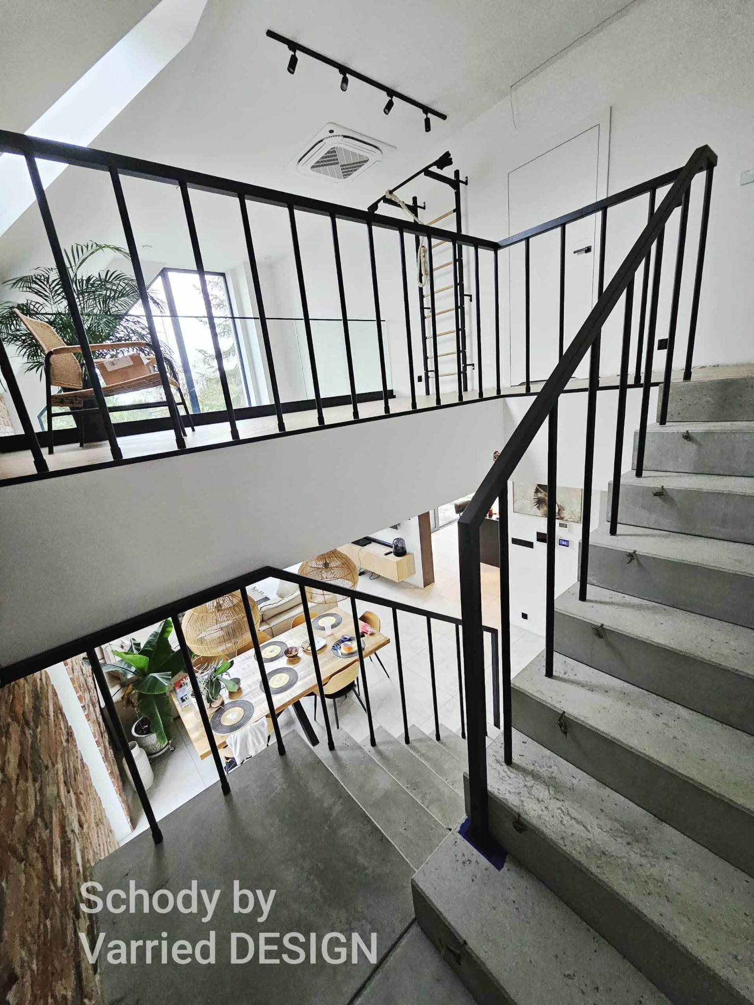 Betonowe schody z czarną, metalową balustradą w nowoczesnym wnętrzu. Widok z góry na stół jadalny i antresolę z roślinami. Minimalistyczny design.