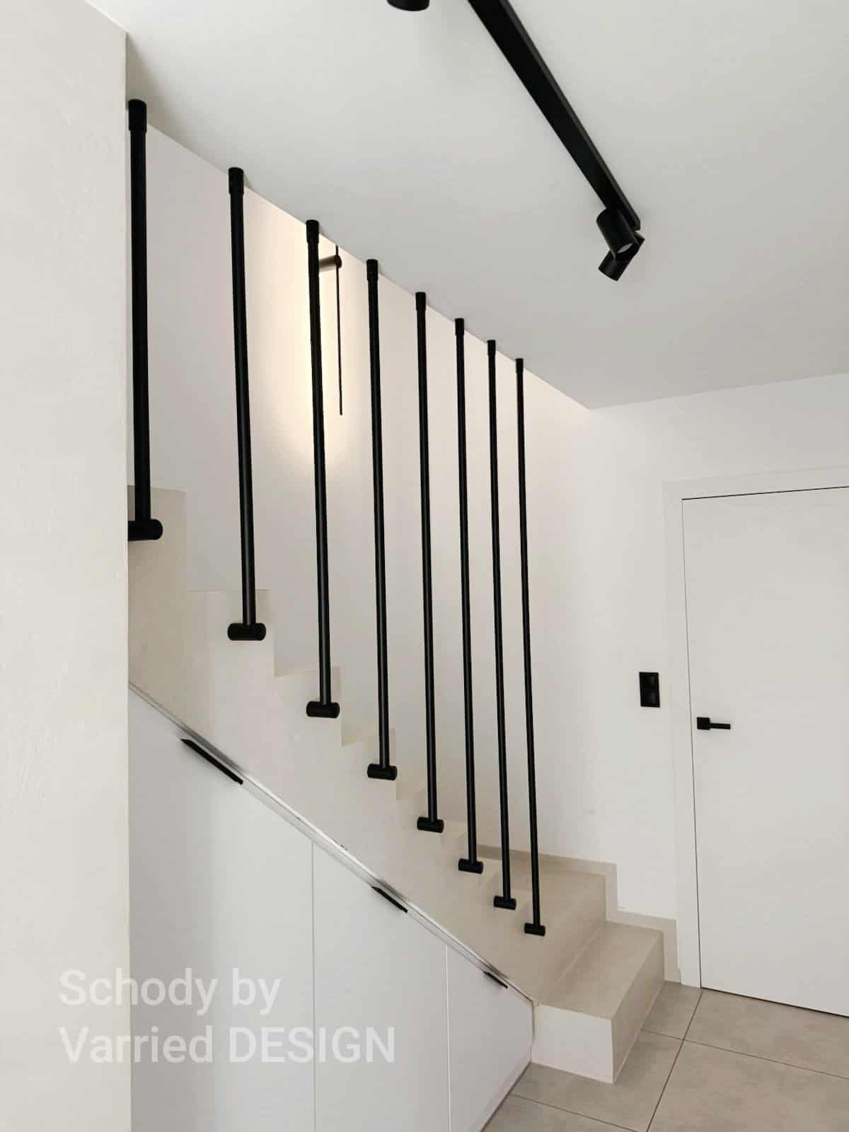 Minimalistyczne schody z czarnymi, pionowymi balustradami na białym tle. Nowoczesny design z oświetleniem punktowym i zabudową pod schodami.