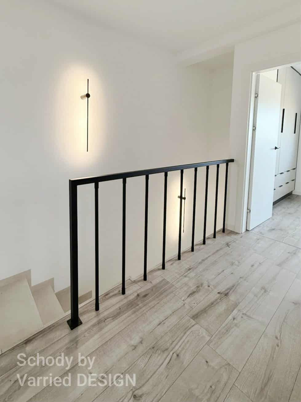 Nowoczesna, czarna poręcz schodowa z minimalistycznym oświetleniem ściennym. Jasne panele podłogowe i białe ściany tworzą eleganckie wnętrze.