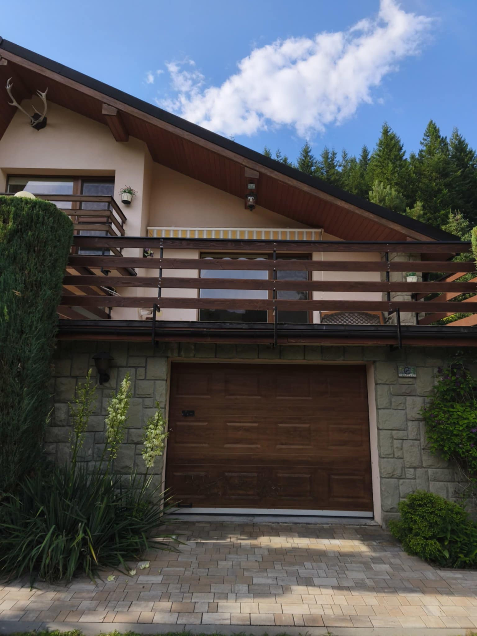 Drewniana balustrada balkonu z poziomymi szczeblami, widok z dołu na tle domu z garażem i zielenią. Uwagę przykuwają detale wykończenia i naturalne materiały.