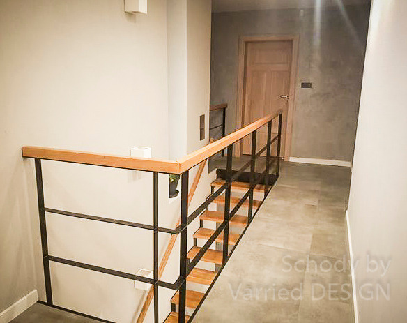 Nowoczesne schody z metalową balustradą i drewnianym poręczą, oświetlone ciepłym światłem, w minimalistycznym wnętrzu z szarą ścianą i drewnianymi drzwiami.