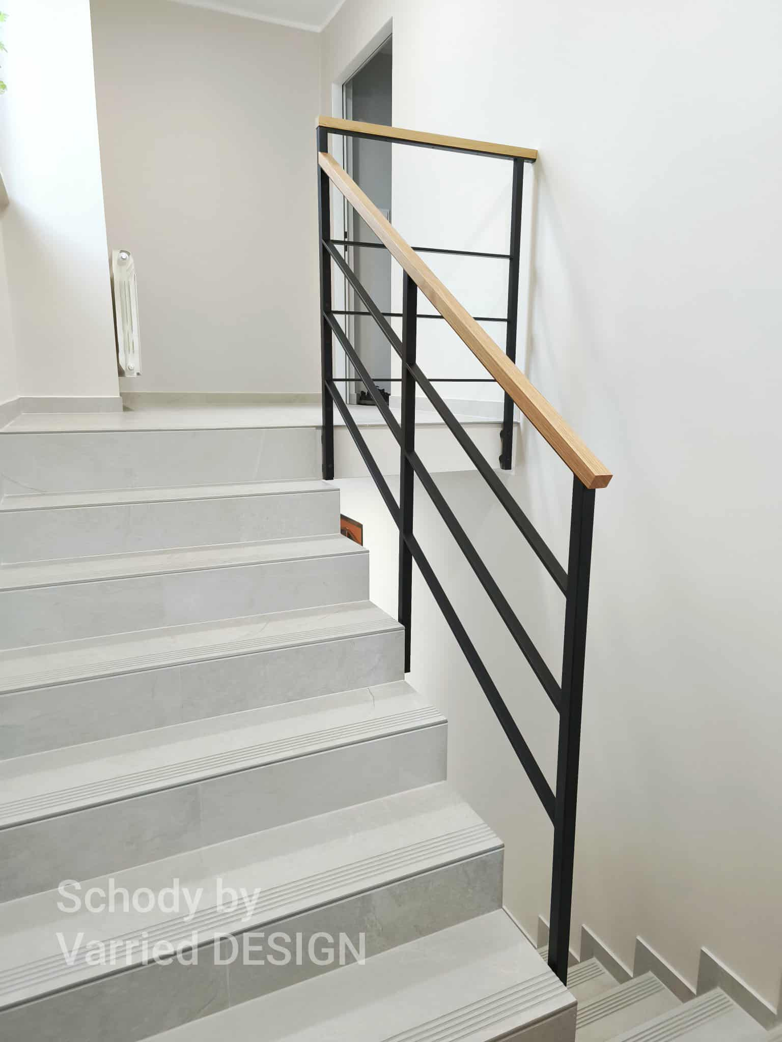 Minimalistyczne schody z szarymi płytkami i czarną, metalową poręczą z drewnianym uchwytem. Ujęcie z góry, podkreślające nowoczesny design i jasne wykończenie wnętrza.