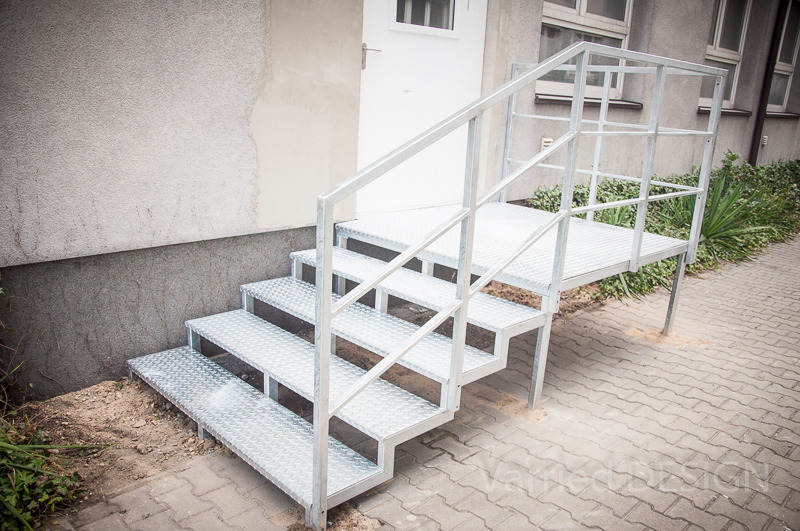 Zewnętrzne schody metalowe z platformą, ocynkowane, z antypoślizgową powierzchnią, prowadzące do białych drzwi wejściowych. Widok z dołu, z boku.