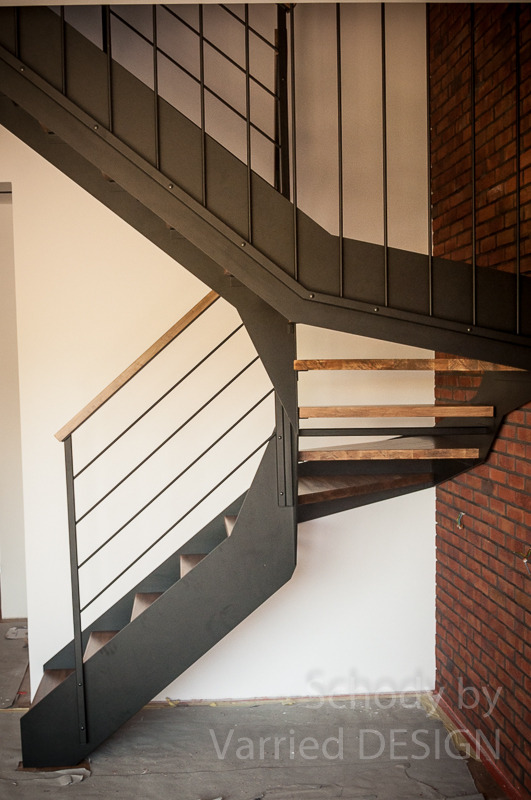 Spiralne schody z metalową konstrukcją i drewnianymi stopniami, z poręczą z jasnego drewna, na tle białej ściany i fragmentu ściany z czerwonej cegły.