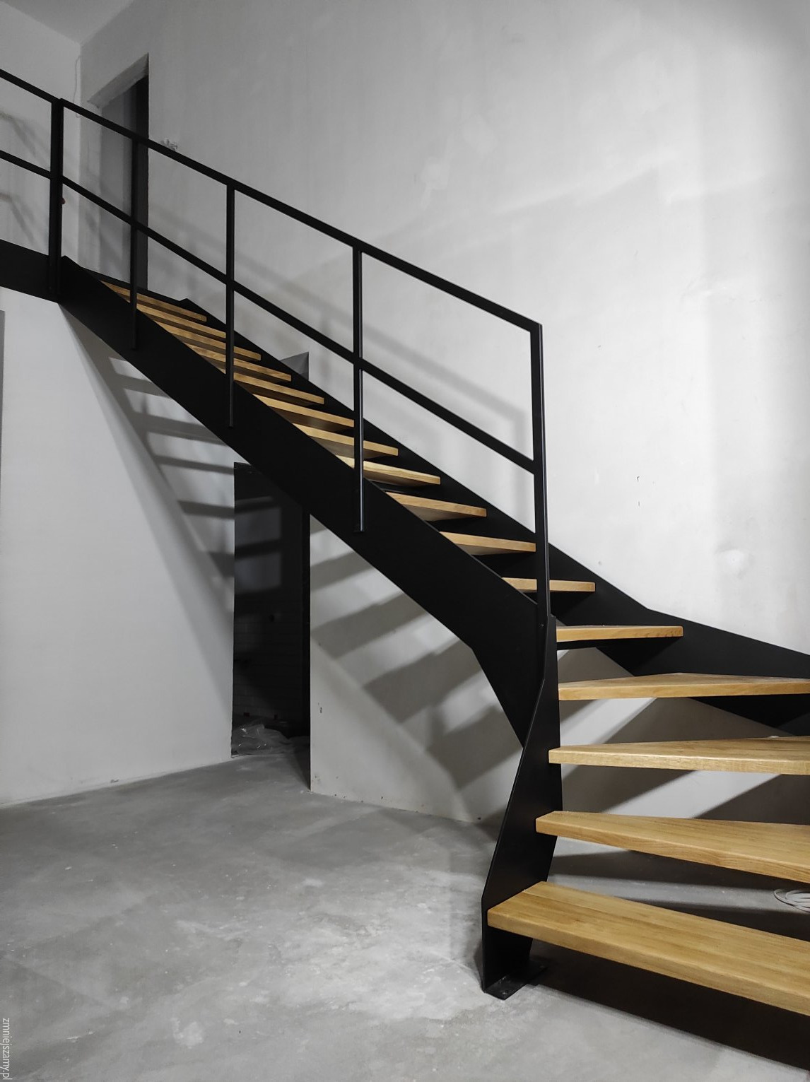 Nowoczesne schody metalowe z drewnianymi stopniami, czarna konstrukcja, minimalistyczny design, widok z dołu, surowe wnętrze, betonowa podłoga.