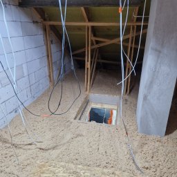 Green Jakub Ogórek - Poddasze w trakcie ocieplania wełną celulozową, widoczne przewody elektryczne z pomarańczowymi znacznikami, konstrukcja drewniana dachu i fragment ściany z bloczków, otwór rewizyjny