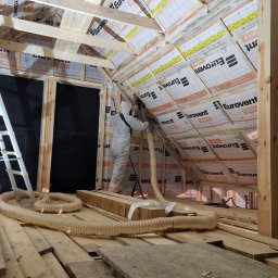 Green Jakub Ogórek - Pracownik w kombinezonie ochronnym aplikuje izolację w przestrzeni poddasza, widoczne drewniane elementy konstrukcji dachu i folia izolacyjna Eurovent.