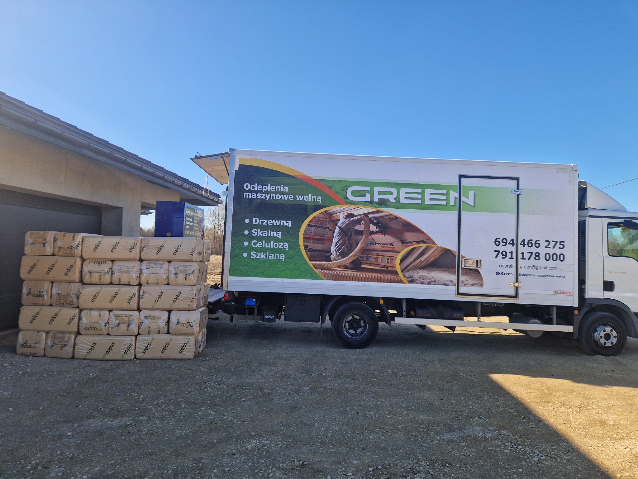 Pakiety izolacji Steico zell ułożone przed budynkiem obok samochodu dostawczego z reklamą ocieplenia maszynowego wełną, w tle błękitne niebo.