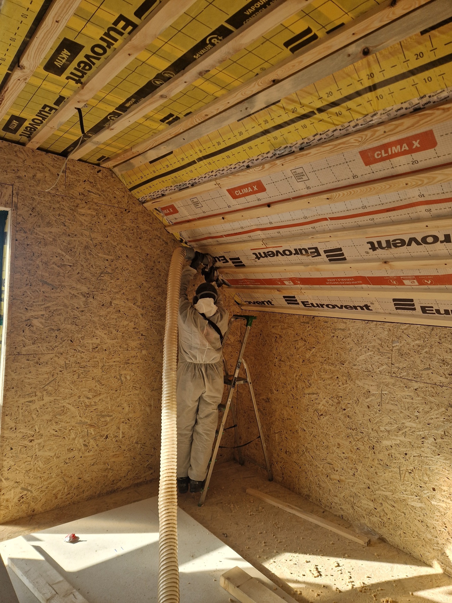 Pracownik w kombinezonie ochronnym na drabinie, w trakcie montażu izolacji poddasza z użyciem węża do wdmuchiwania materiału izolacyjnego; widoczne drewniane krokwie i płyty OSB.