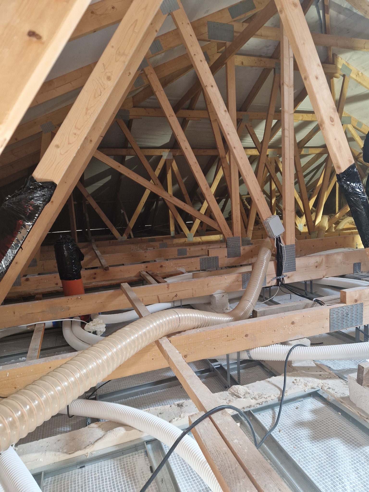 Konstrukcja drewniana poddasza z widocznymi elementami instalacji wentylacyjnej i izolacji, z detalami takimi jak metalowe łączniki i folia zabezpieczająca.
