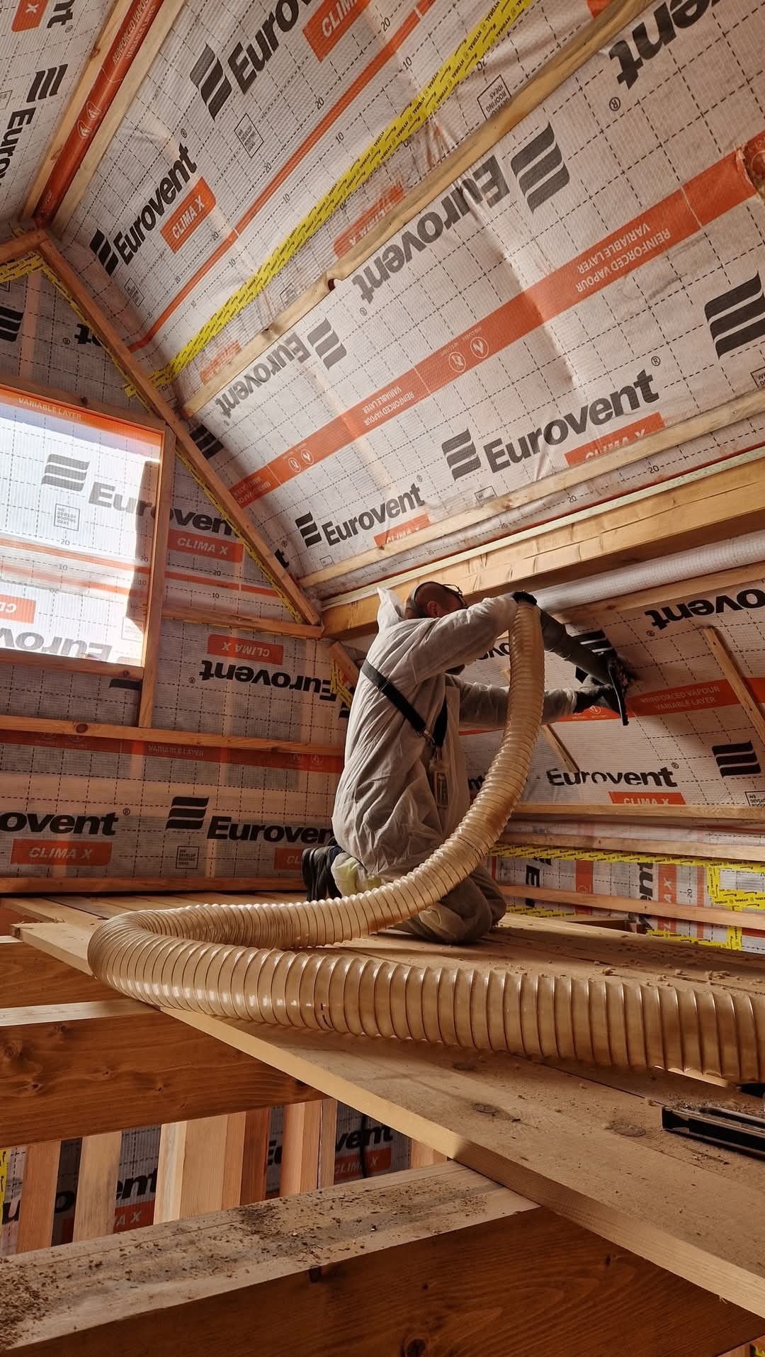 Pracownik w kombinezonie ochronnym aplikuje izolację natryskową na poddaszu w Platerowie, widoczny duży wąż doprowadzający materiał izolacyjny i folia Eurovent na krokwiach.