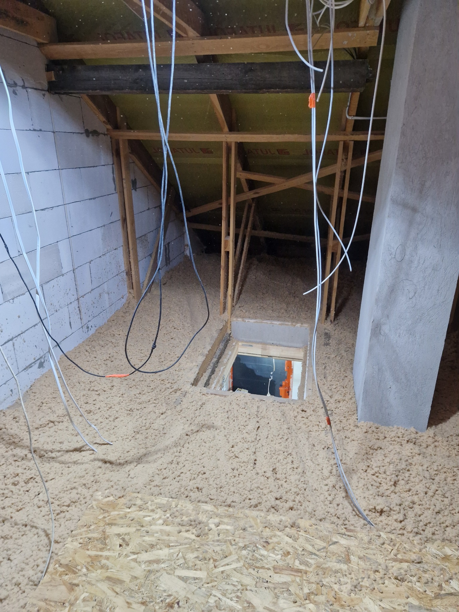 Poddasze w trakcie ocieplania wełną celulozową, widoczne przewody elektryczne z pomarańczowymi znacznikami, konstrukcja drewniana dachu i fragment ściany z bloczków, otwór rewizyjny