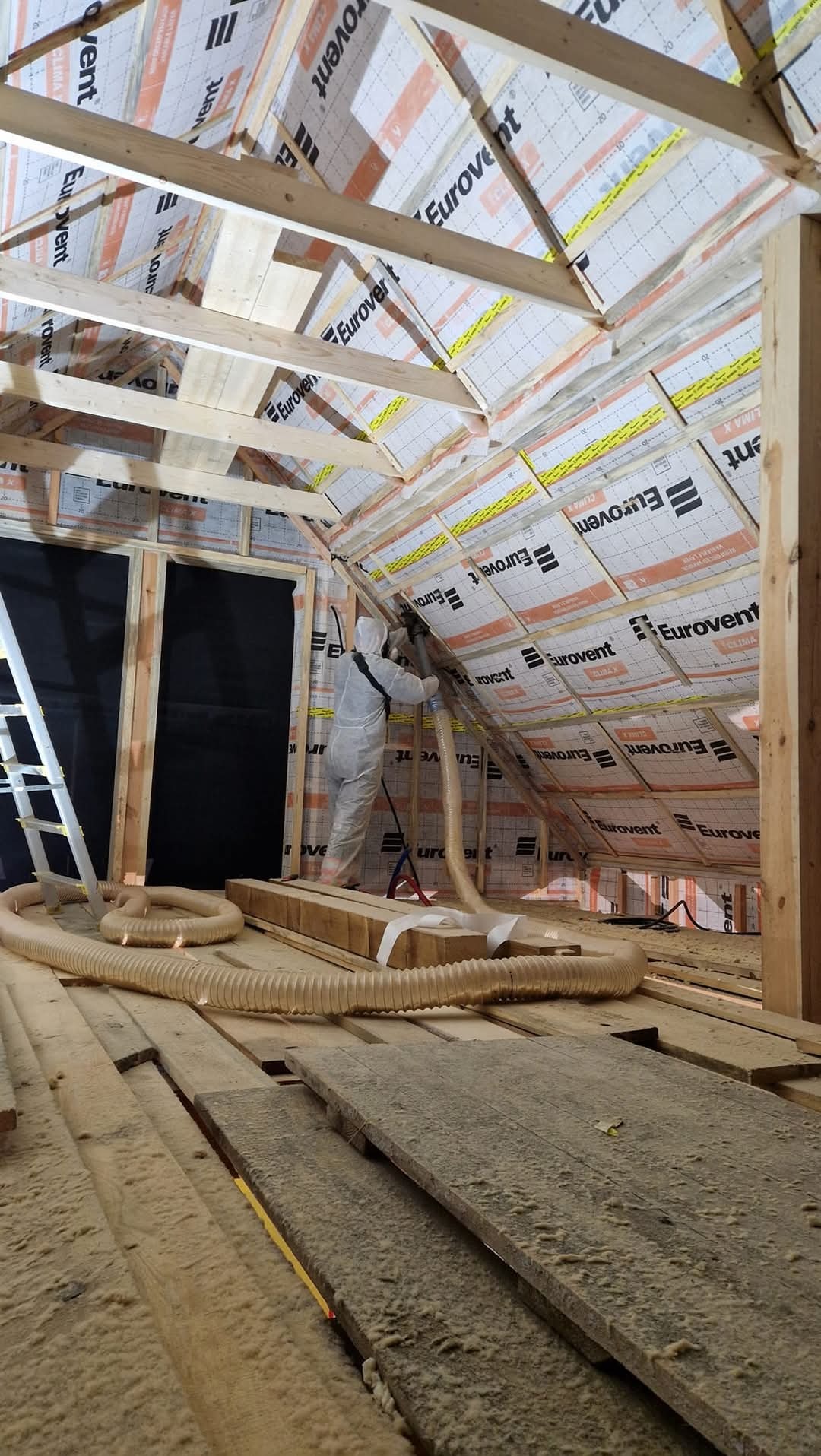 Pracownik w kombinezonie ochronnym aplikuje izolację w przestrzeni poddasza, widoczne drewniane elementy konstrukcji dachu i folia izolacyjna Eurovent.
