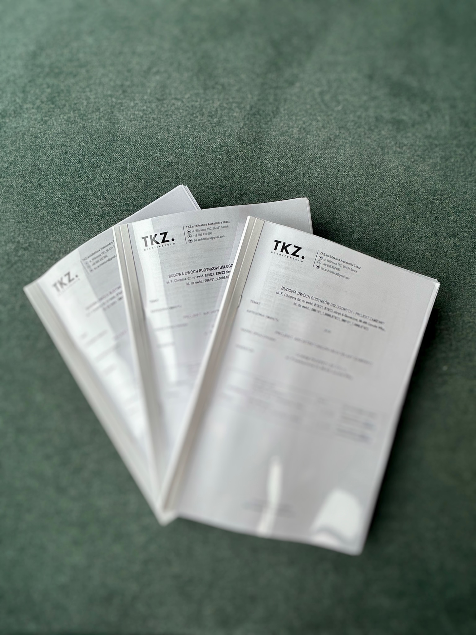 Cztery białe teczki z logo TKZ architektura Aleksandra Tkacz, ułożone ukośnie na zielonym tle, prezentujące projekt budowy dwóch budynków usługowych.