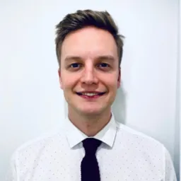 Uśmiechnięty młody mężczyzna w białej koszuli w czarne kropki i bordowym krawacie na tle białej ściany.