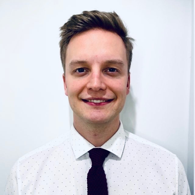 Uśmiechnięty młody mężczyzna w białej koszuli w czarne kropki i bordowym krawacie na tle białej ściany.