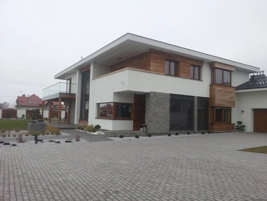 Nowoczesny dom z białą elewacją, drewnianymi akcentami i kamienną okładziną. Widoczny balkon z szklaną balustradą i podjazd z kostki brukowej.