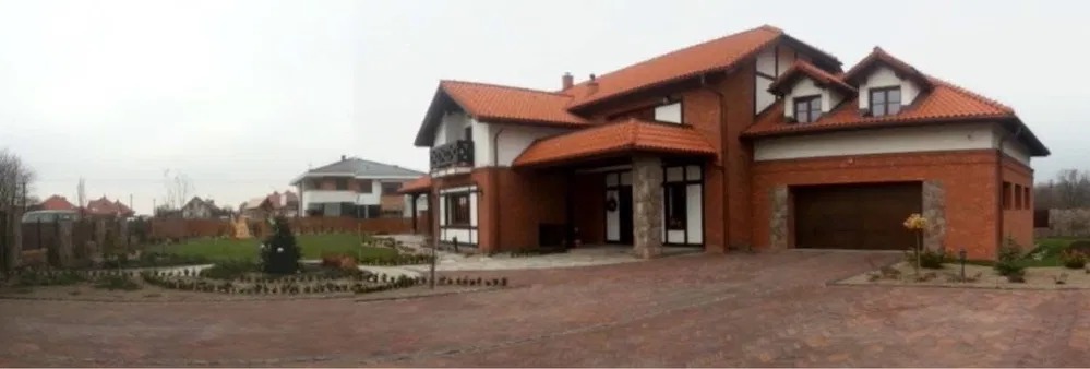 Dwukondygnacyjny dom murowany z czerwoną dachówką i garażem, otoczony ogrodem z kostki brukowej, widok z szerokiego kąta w pochmurny dzień.