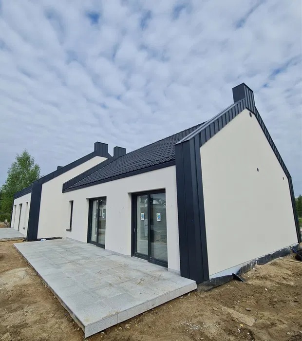 Nowoczesny dom z białą elewacją i ciemnymi elementami wykończeniowymi, widok z zewnątrz, betonowy taras, dachówka w kolorze grafitu, błękitne niebo.