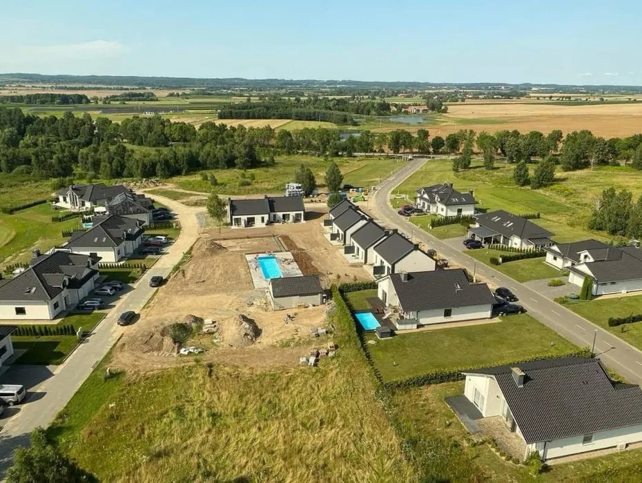 Osiedle domów jednorodzinnych z ciemnymi dachami, widok z lotu ptaka. Jeden z domów w trakcie budowy, z widocznym basenem. Słoneczny dzień, zielone pola w tle.