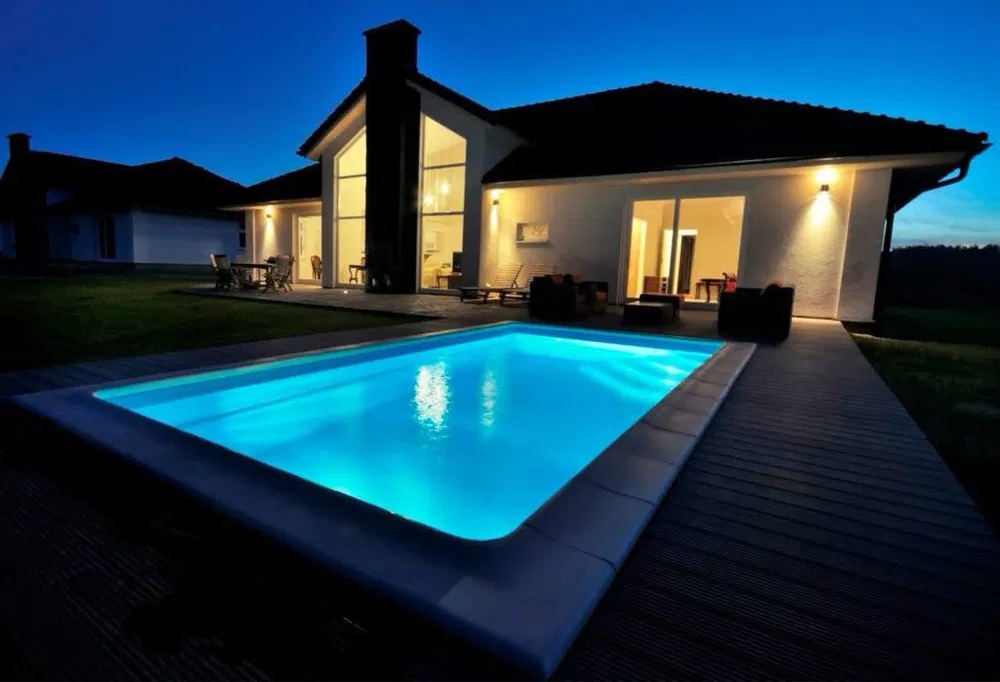 Nowoczesny dom z basenem oświetlonym nocą. Biała elewacja, duże okna i drewniany taras tworzą elegancki wygląd. Widoczne oświetlenie zewnętrzne.