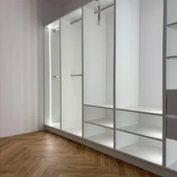 Biała, otwarta szafa wnękowa z półkami i drążkami na ubrania, oświetlona LED, na tle parkietu ułożonego w jodełkę, w jasnym wnętrzu.