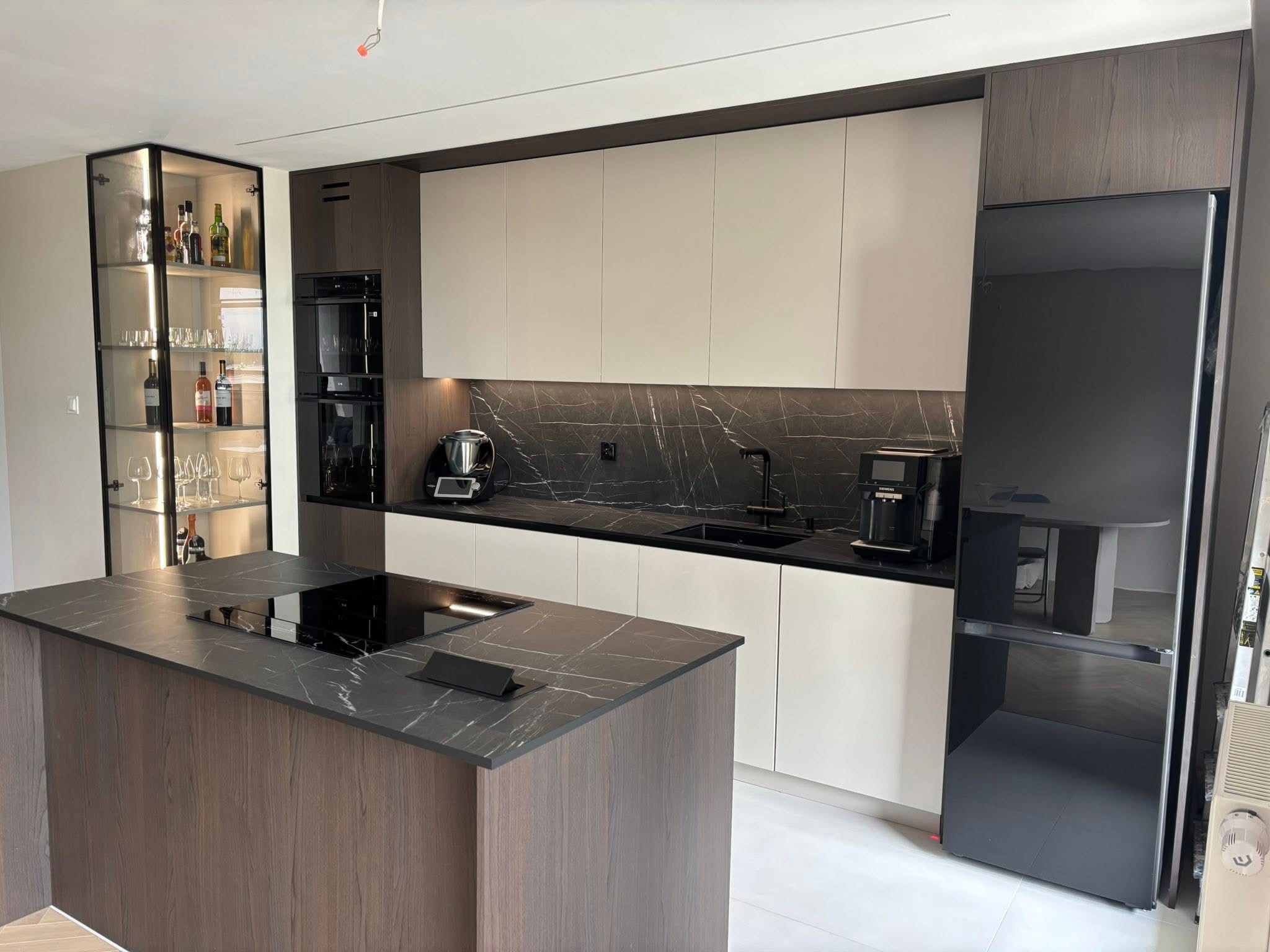 Nowoczesna kuchnia z wyspą i czarnym blatem imitującym marmur. Szklana witryna z oświetleniem eksponuje szkło i alkohole. Minimalistyczny design.