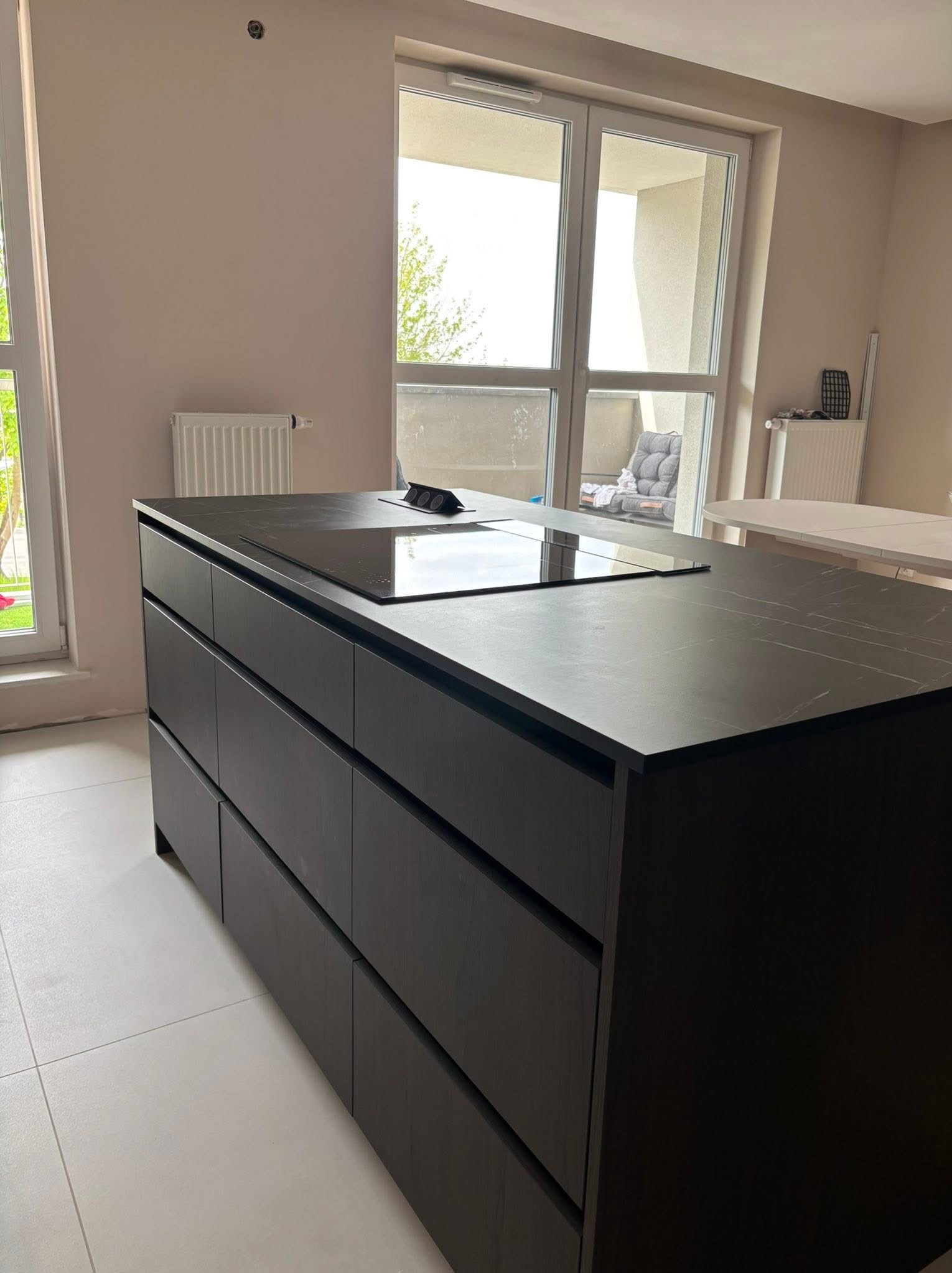 Nowoczesna, ciemna wyspa kuchenna z płytą indukcyjną i marmurowym blatem, na tle jasnego wnętrza z dużym oknem balkonowym i grzejnikiem.