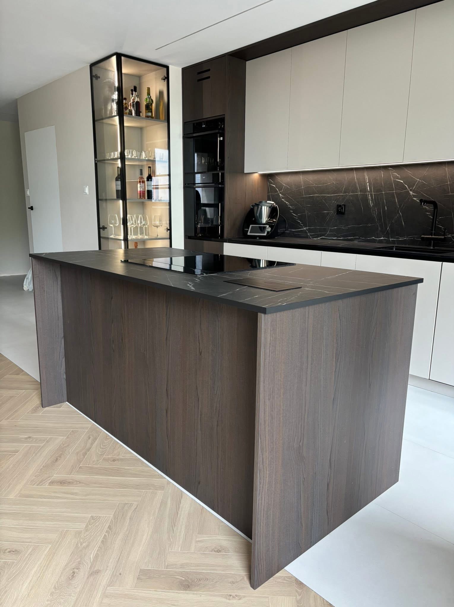 Nowoczesna kuchnia z wyspą, fronty w odcieniach brązu i bieli, czarny blat. Na wyspie płyta indukcyjna. Szklana witryna z oświetleniem i alkoholem w tle.