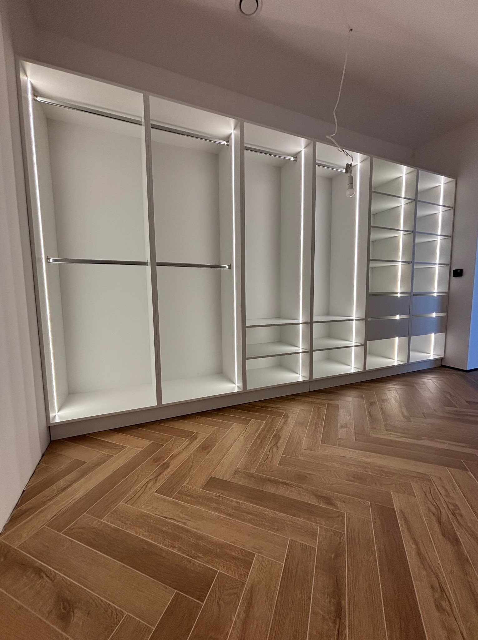 Biała szafa wnękowa z oświetleniem LED w Elblągu. Widoczny parkiet ułożony w jodełkę i niedokończone oświetlenie sufitowe. Szafa posiada półki i drążki.