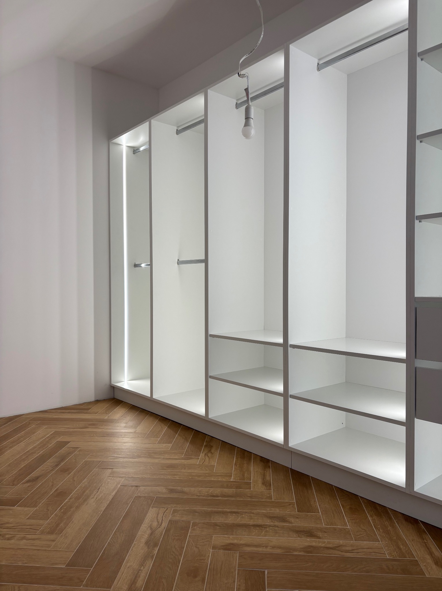 Biała, otwarta szafa wnękowa z półkami i drążkami na ubrania, oświetlona LED, na tle parkietu ułożonego w jodełkę, w jasnym wnętrzu.