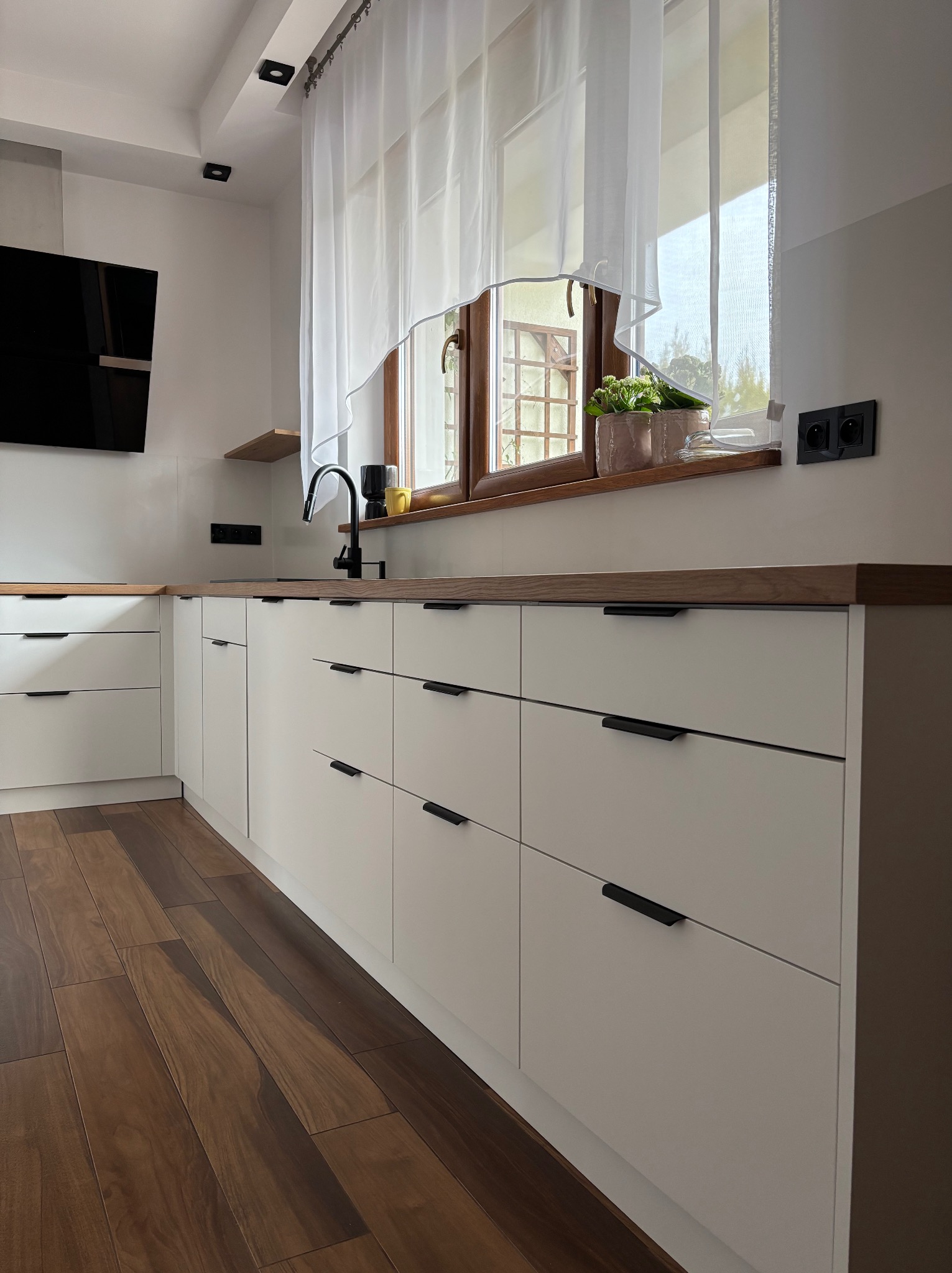 Jasna kuchnia z drewnianym blatem i podłogą. Białe fronty szafek z czarnymi, podłużnymi uchwytami. Okno z firanką i roślinami. Nowoczesny design.
