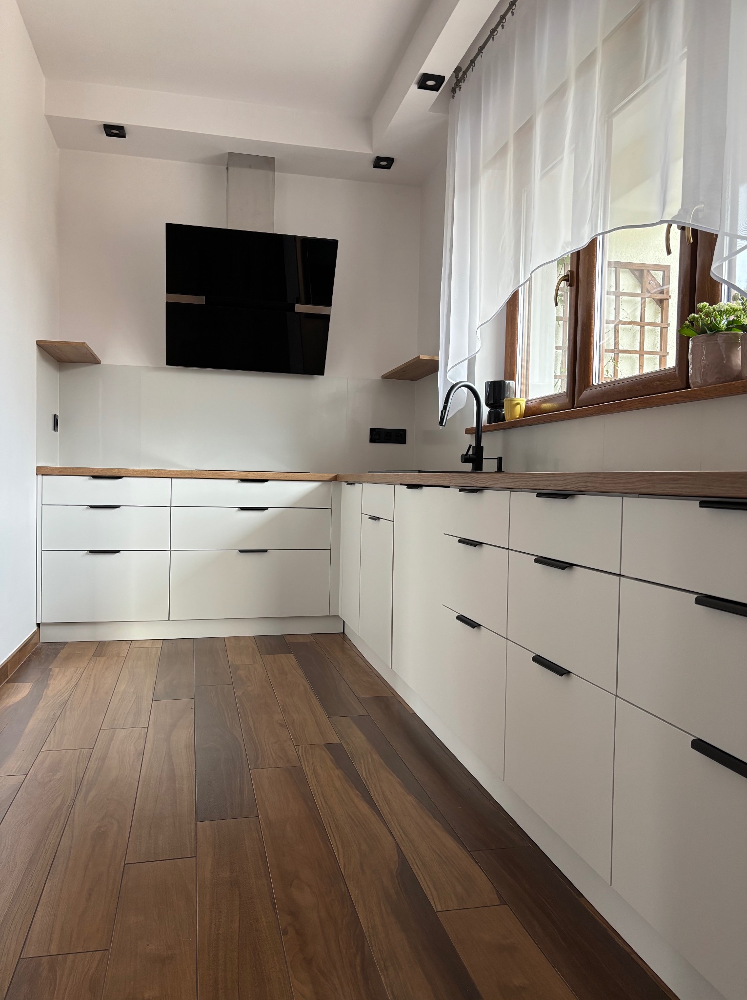 Jasna kuchnia z drewnianym blatem i czarnymi detalami: okap, bateria, uchwyty. Uwagę przykuwa drewniana podłoga i rośliny w oknie.