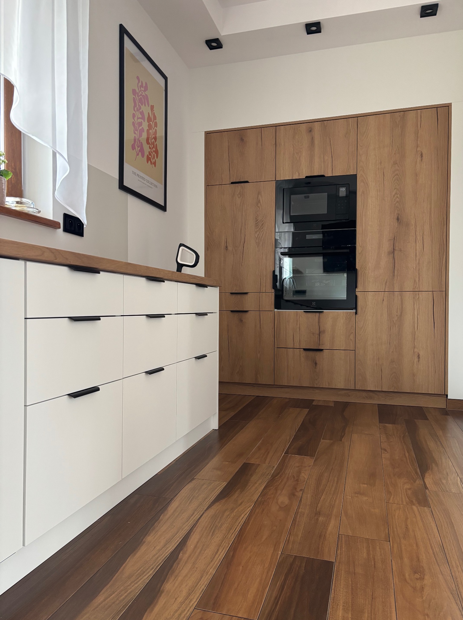 Nowoczesna kuchnia: białe fronty szafek z drewnianym blatem i zabudowa z forniru w odcieniach drewna, czarne uchwyty, parkiet w jodełkę, plakat na ścianie.