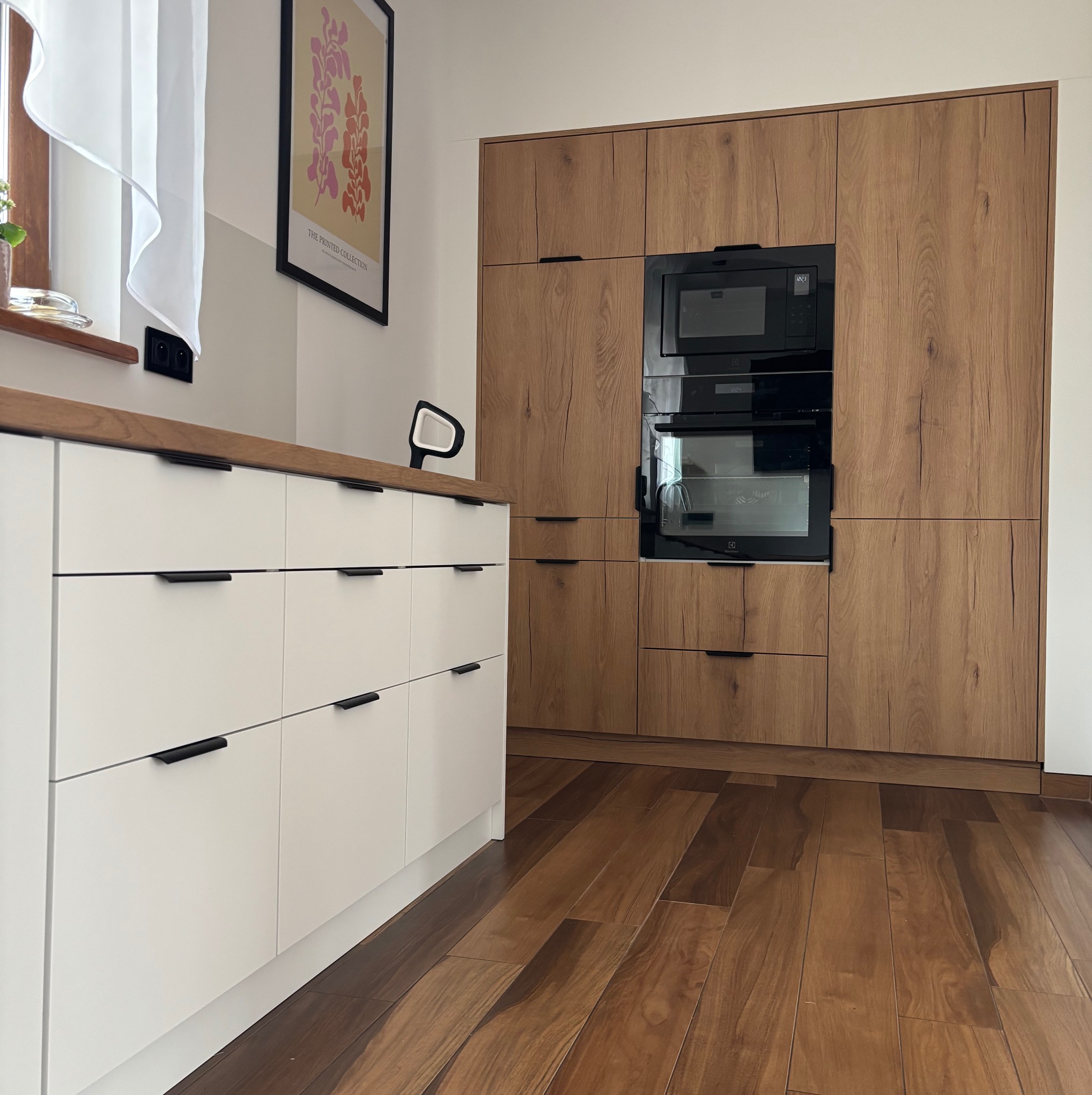 Nowoczesna kuchnia: białe szafki z czarnymi uchwytami i drewnopodobna zabudowa z piekarnikiem i mikrofalówką, parkiet. Minimalistyczny design.