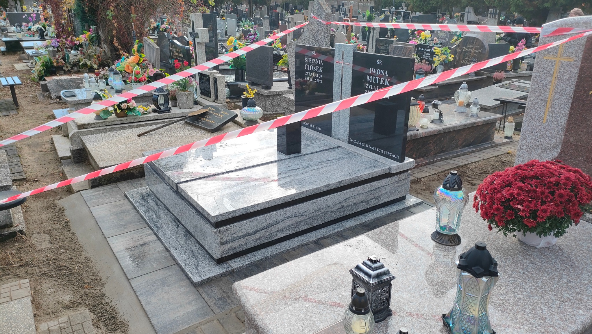 Nowy, dwupoziomowy nagrobek z szarego granitu, otoczony taśmą ostrzegawczą na cmentarzu. Widoczne lampiony i kwiaty. Nagrobek z czarną tablicą inskrypcyjną.