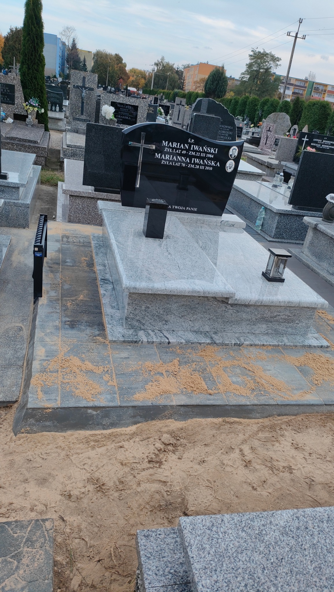 Granitowy nagrobek z czarną tablicą i krzyżem na cmentarzu, otoczony piaskiem. Widoczne inne nagrobki i budynki w tle. Ujęcie z dołu.