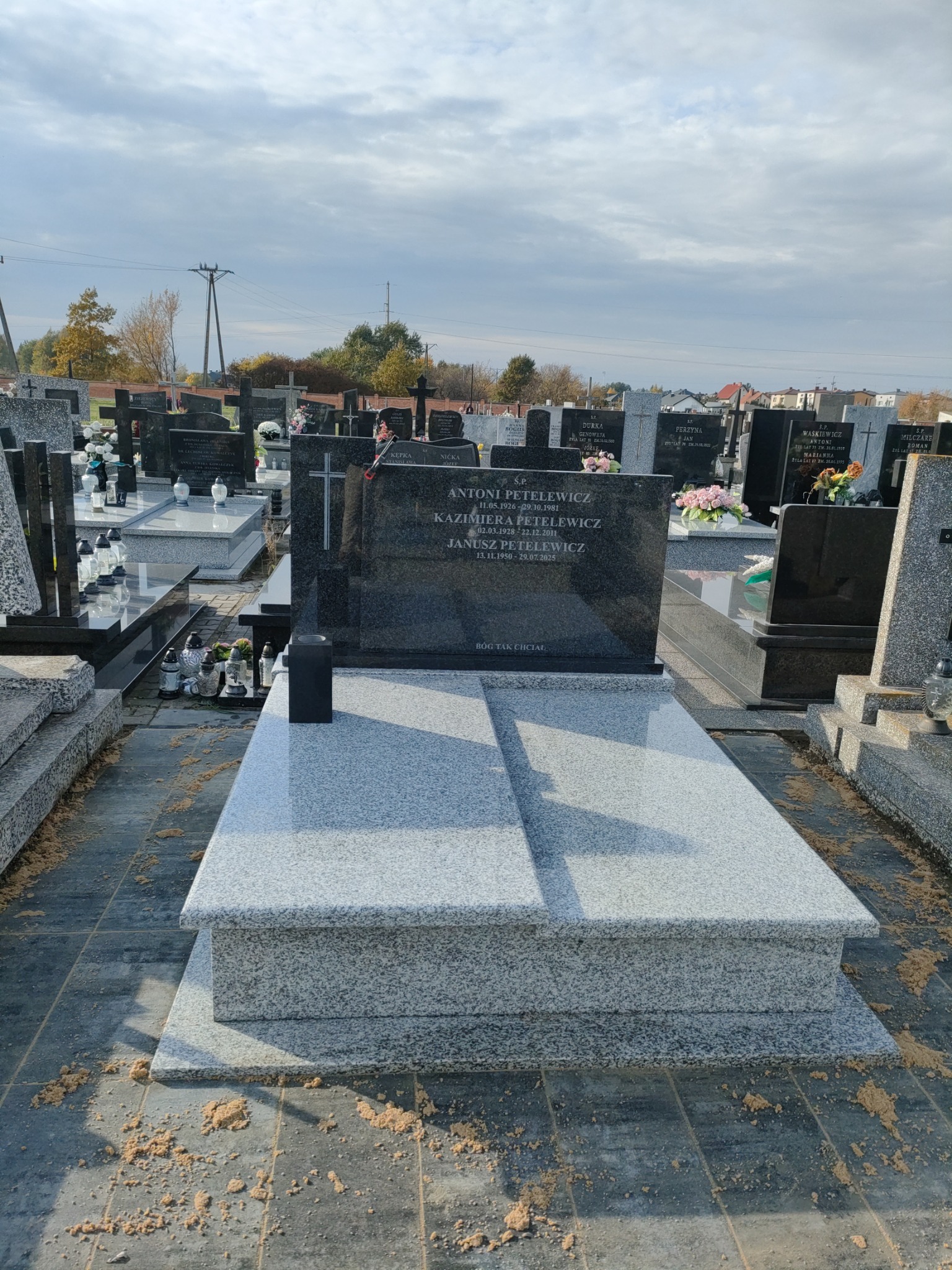 Granitowy nagrobek z inskrypcjami na cmentarzu w Łodzi, otoczony innymi nagrobkami. Widok na pochmurne niebo i drzewa w tle. Detal kamieniarski.