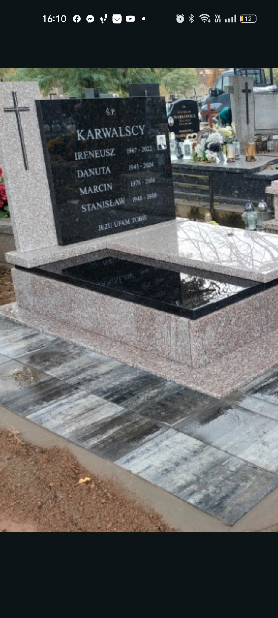Czarny nagrobek z jasnoszarym obramowaniem i krzyżem, z inskrypcjami i zdjęciem zmarłych, otoczony szarą kostką brukową na cmentarzu.