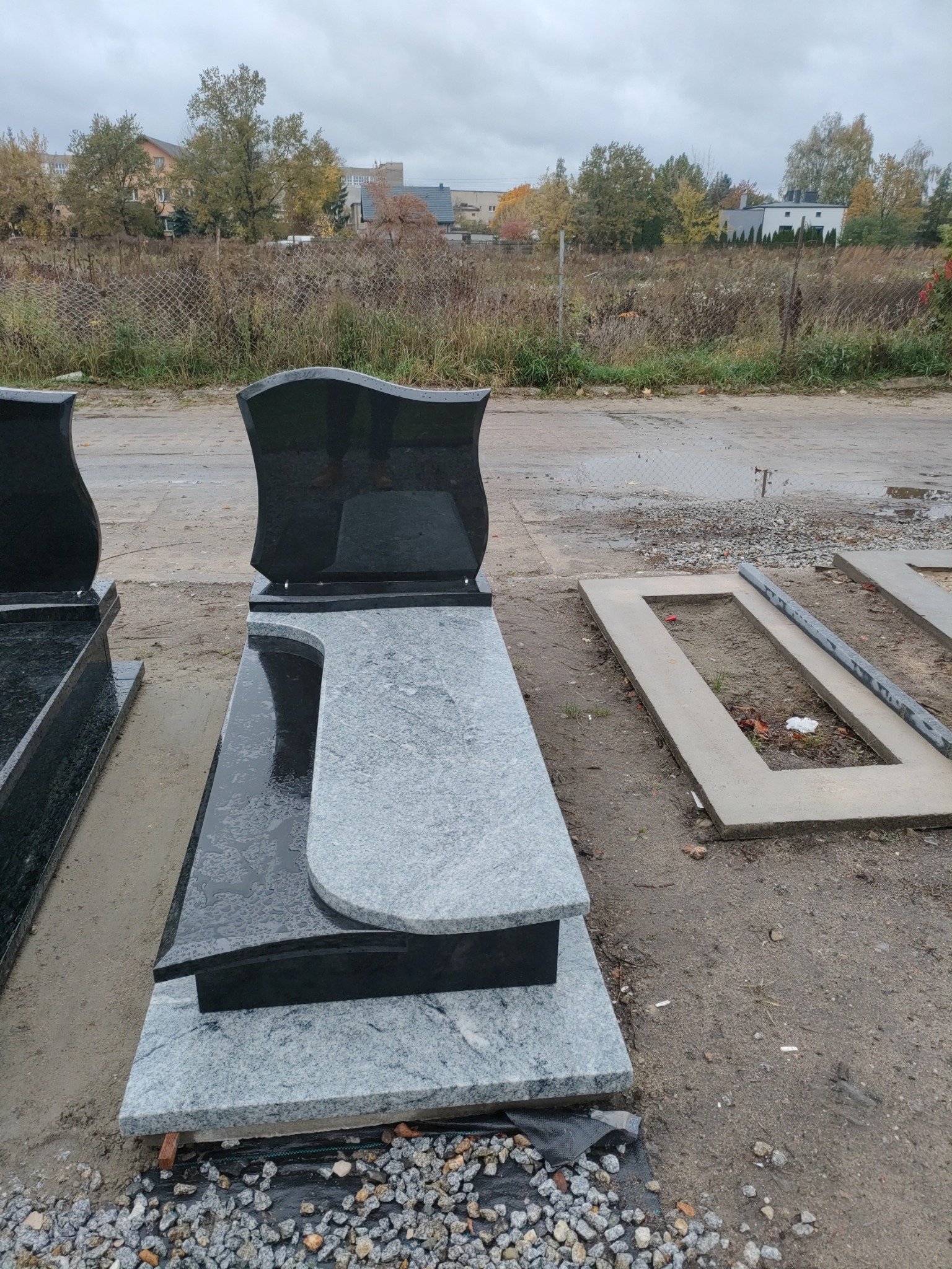 Nowy, dwukolorowy nagrobek z granitu, z elementami w kolorze czarnym i szarym, ustawiony na placu kamieniarskim. W tle widoczne drzewa i budynki.