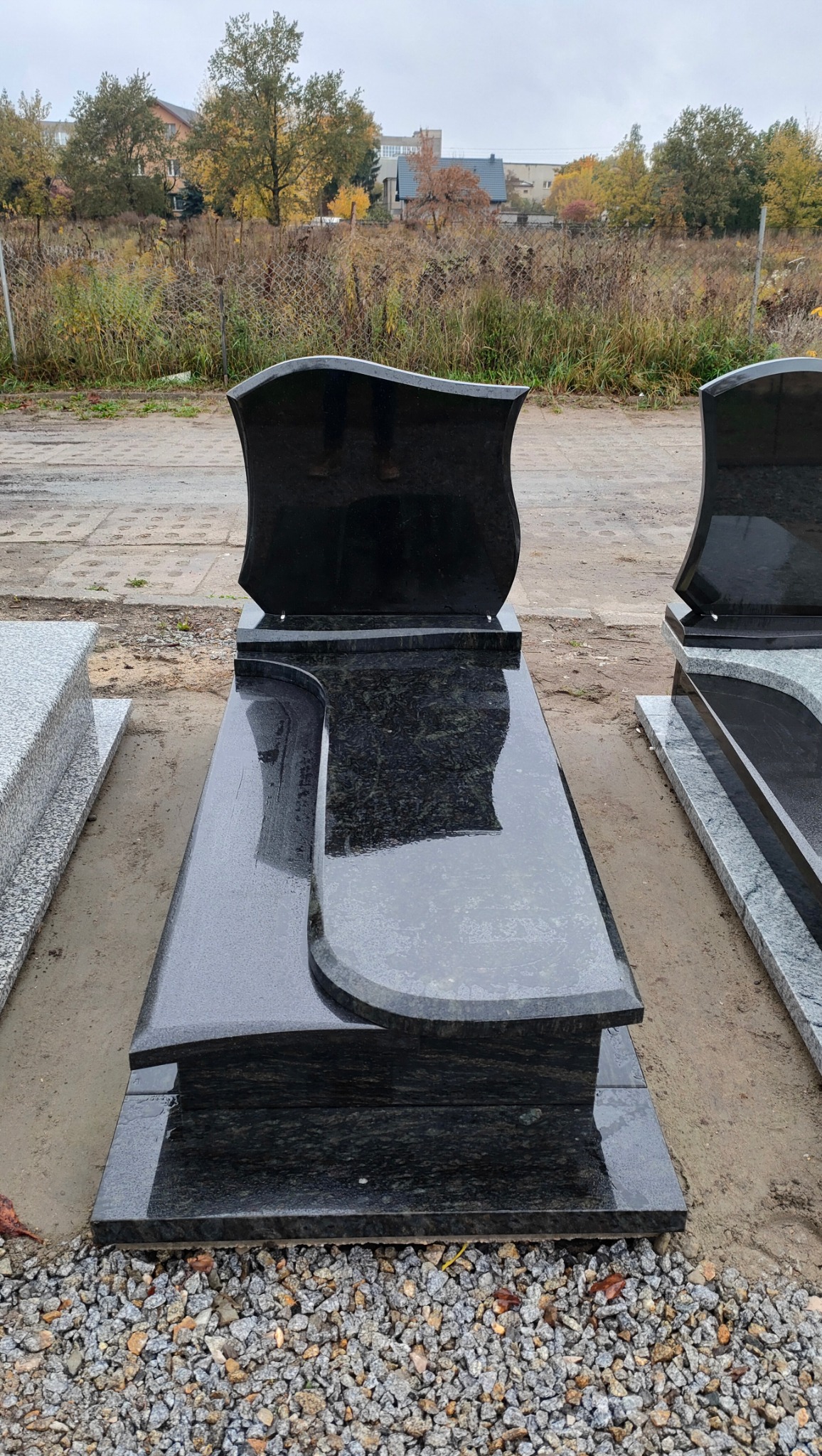 Elegancki, czarny nagrobek z granitu o falistym kształcie, stojący na żwirowym podłożu. W tle widoczne inne nagrobki i zieleń.