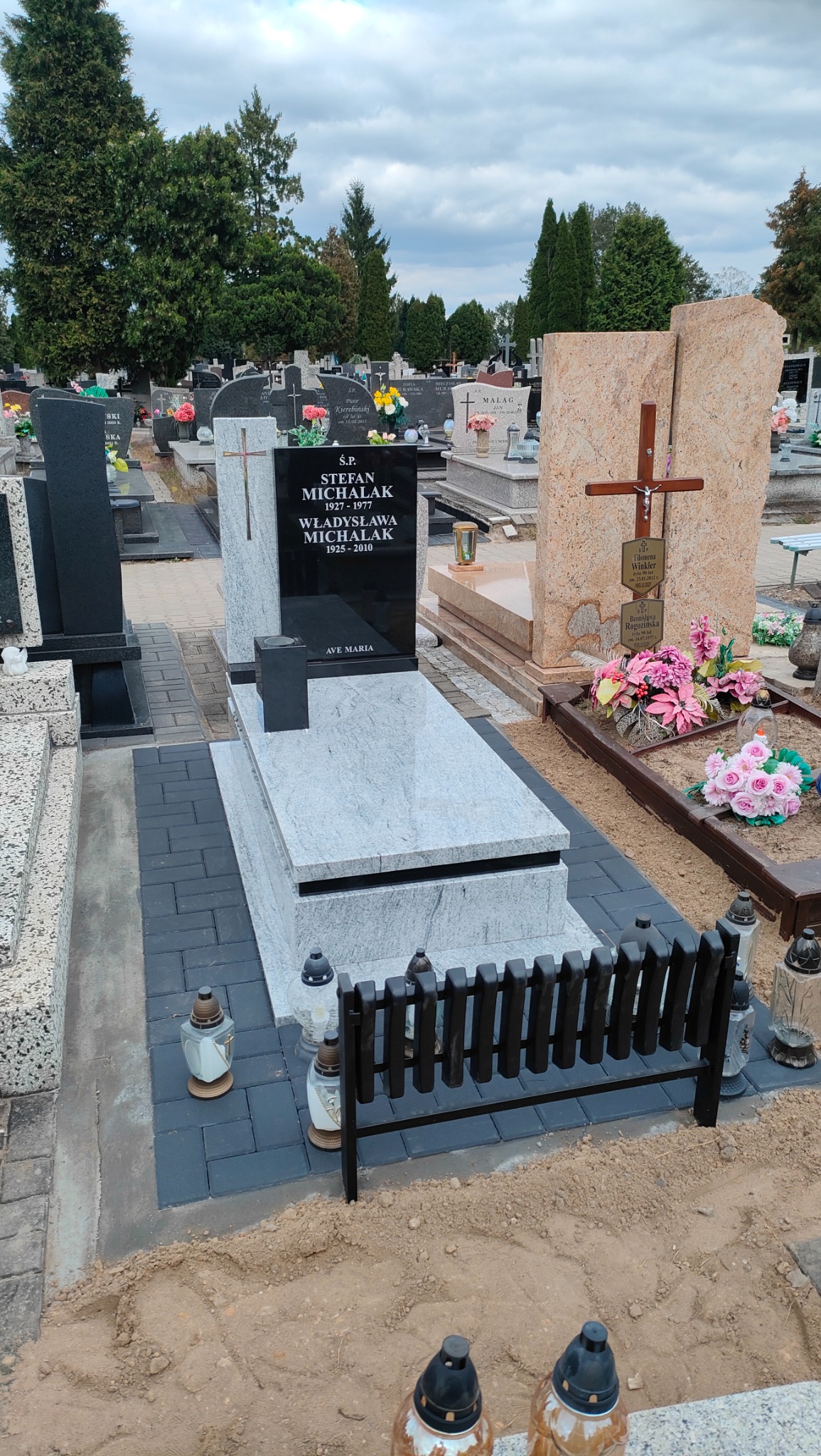 Granitowy nagrobek z czarną tablicą inskrypcyjną, otoczony szarą kostką brukową i drewnianym płotkiem. Widok na cmentarz z innymi nagrobkami i drzewami w tle.