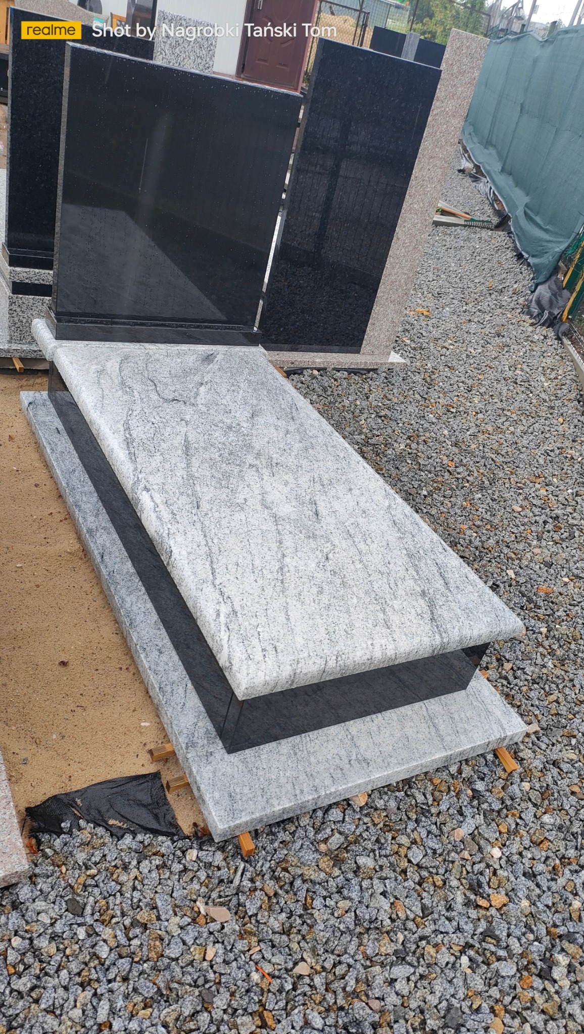 Jasnoszary nagrobek z ciemnym elementem, ustawiony na żwirowym podłożu. Widoczne inne nagrobki w tle. Ujęcie z góry, podkreślające fakturę kamienia.